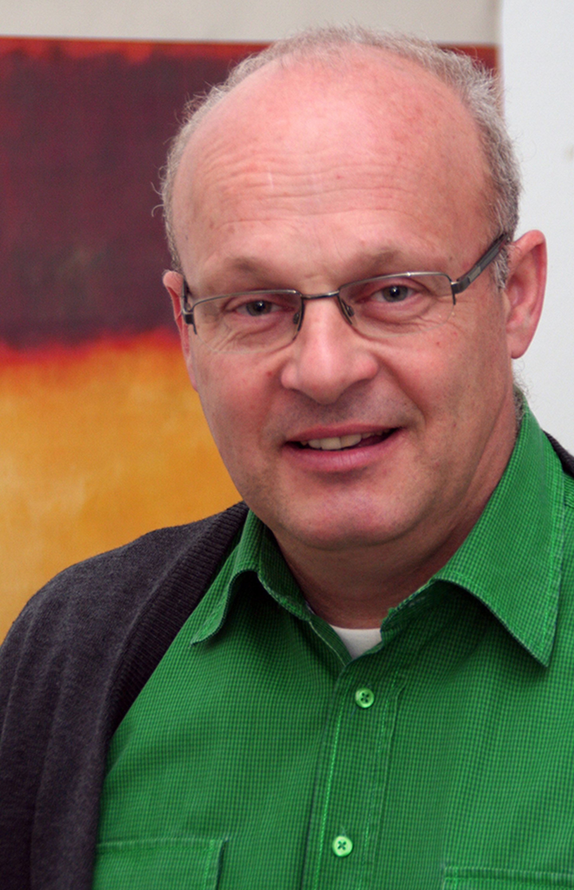 Lächelnder Mann mit grauen Haaren und Brille, der ein grünes Hemd mit einer dunkelgrauen Jacke trägt und vor einem Gemälde steht