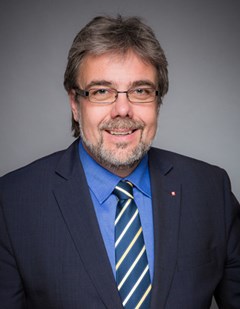 Lächelnder Mann mit grauen Haaren und Bart, der ein blaues Hemd, eine blau-gelb-gestreifte Krawatte und eine schwarze Anzugjacke trägt