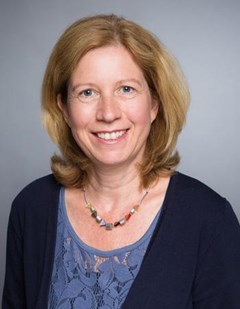 Lächelnde Frau mit schulterlangen, blonden Haaren, die ein hellblaues Shirt, eine dunkelblaue Jacke und eine bunte Halskette trägt