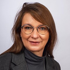 Lächelnde Frau mit langen, braunen Haaren und Brille, die schwarze Oberbekleidung trägt